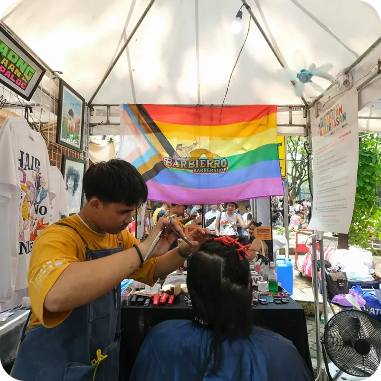 Barber cutting hair at a Pride-themed pop-up shop with a rainbow flag and Barbierro Barbershop sign