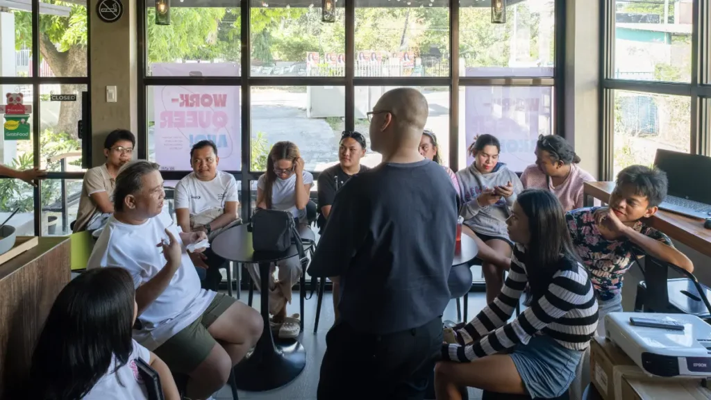 A speaker presenting to a group of people gathered around a small table inside a cafe space.